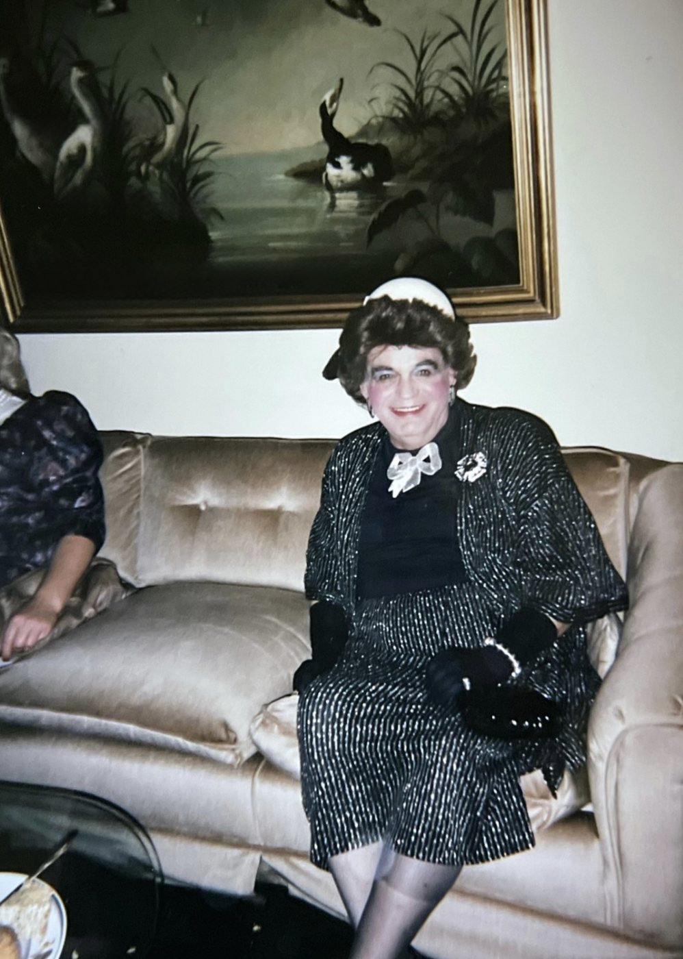Uncle Walter in his drag persona Lottie — pillbox hat and beaded black dress, seated on a tufted gold velvet sofa beneath a vintage painting of swans